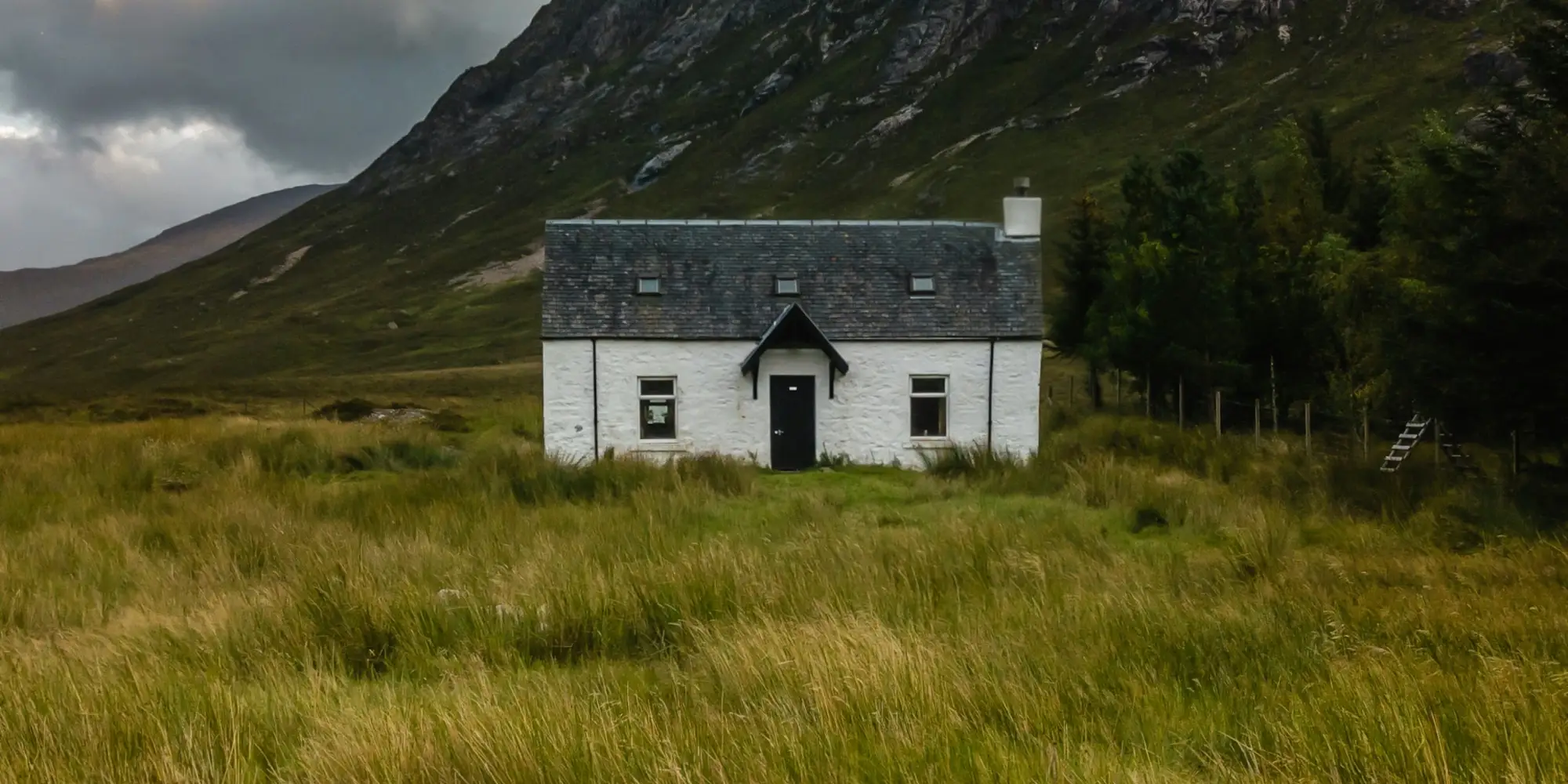 Weißes Cottage im Gras vor bewaldeten Berghängen in einem schottischen Tal